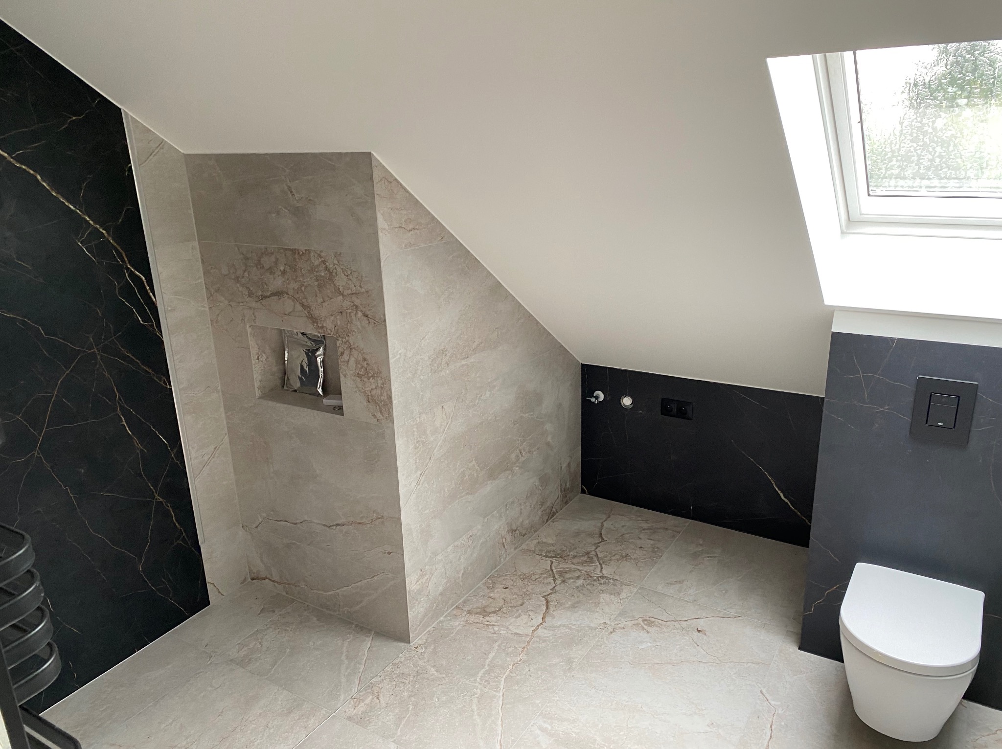 Nowoczesna łazienka na poddaszu z czarnymi i beżowymi płytkami imitującymi marmur, biała wisząca toaleta i okno dachowe.