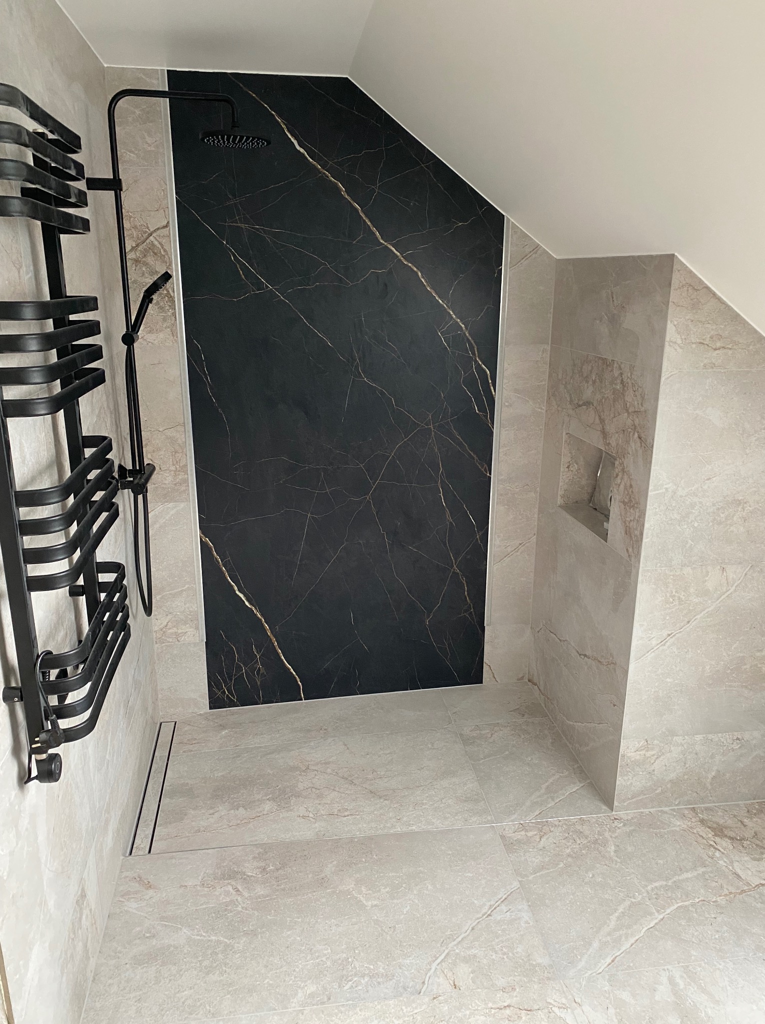 Wykończona łazienka na poddaszu z czarną ścianą prysznicową imitującą marmur, beżowe płytki na podłodze i ścianach, odpływ liniowy w podłodze, czarny grzejnik drabinkowy i wnęka w ścianie.