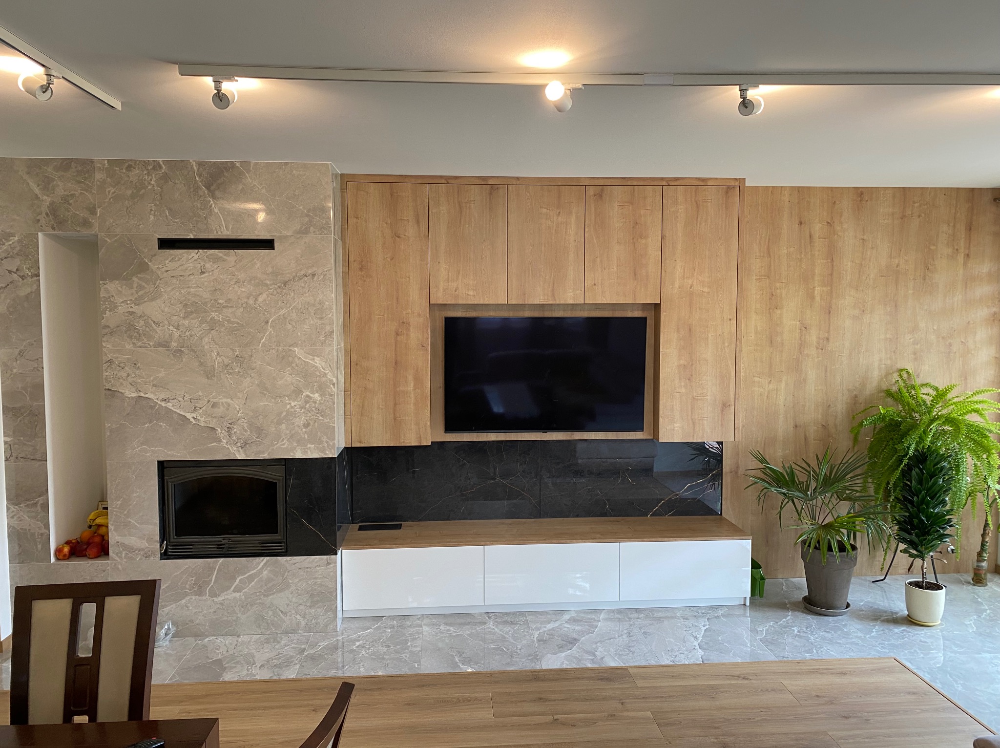 Salon z marmurowym kominkiem, zabudową meblową z drewnianymi frontami, telewizorem i roślinami doniczkowymi, wykończony w szarościach i drewnie.