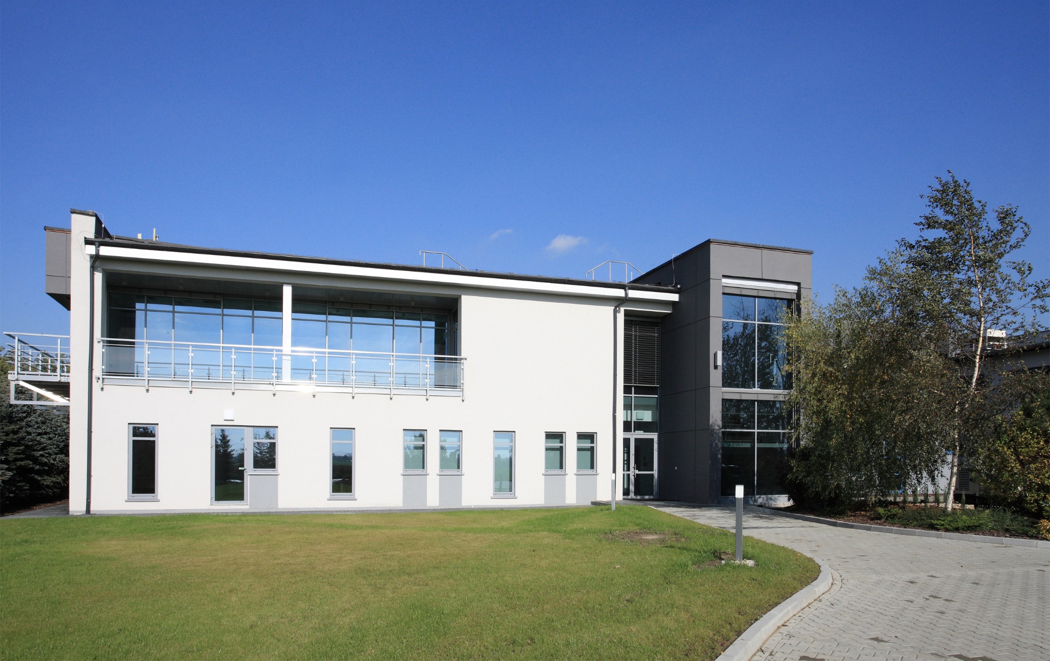 Dwukondygnacyjny budynek usługowy z białymi i szarymi elewacjami, dużymi oknami i metalowymi balustradami, otoczony trawnikiem i drzewami, widok z poziomu gruntu w słoneczny dzień.