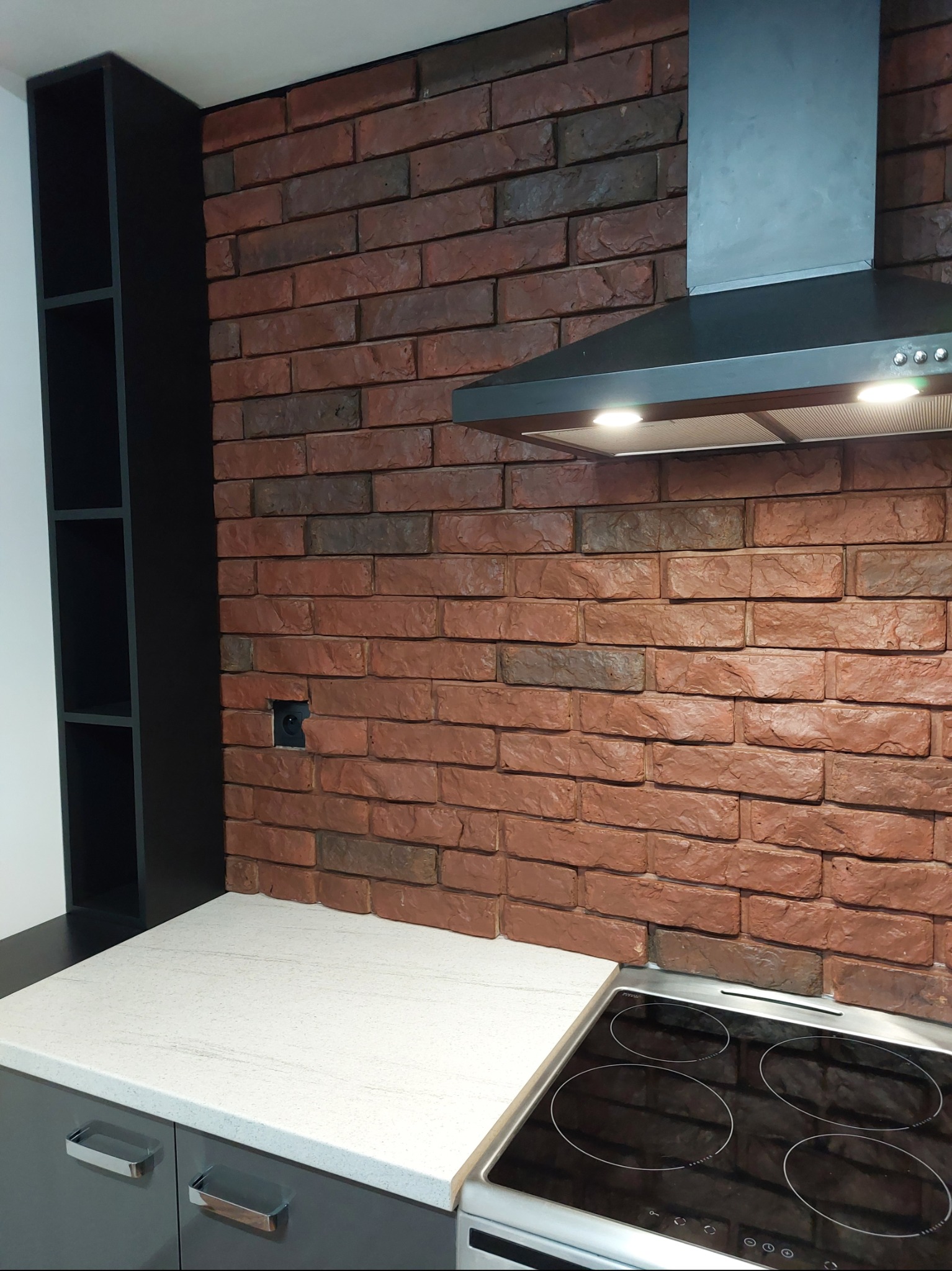 Ceglana ściana w kuchni z czarnym okapem i szafką. Widoczna płyta indukcyjna i blat. Dekoracyjna ściana z imitacji cegły w odcieniach brązu i szarości.
