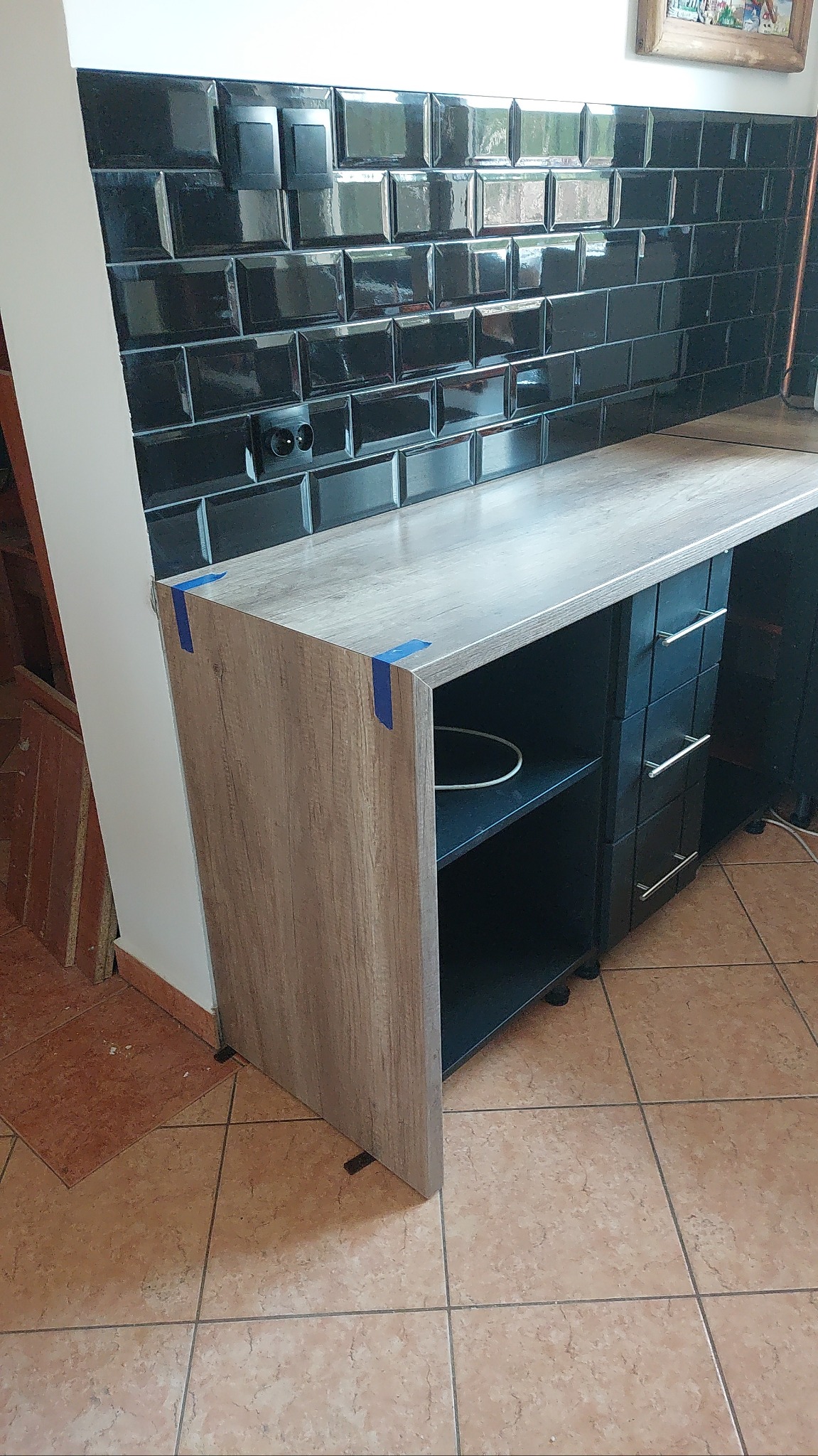 Częściowy widok blatu kuchennego z czarnymi płytkami na ścianie, zabezpieczonego niebieską taśmą. Widoczne szuflady i półki. Na podłodze płytki ceramiczne.