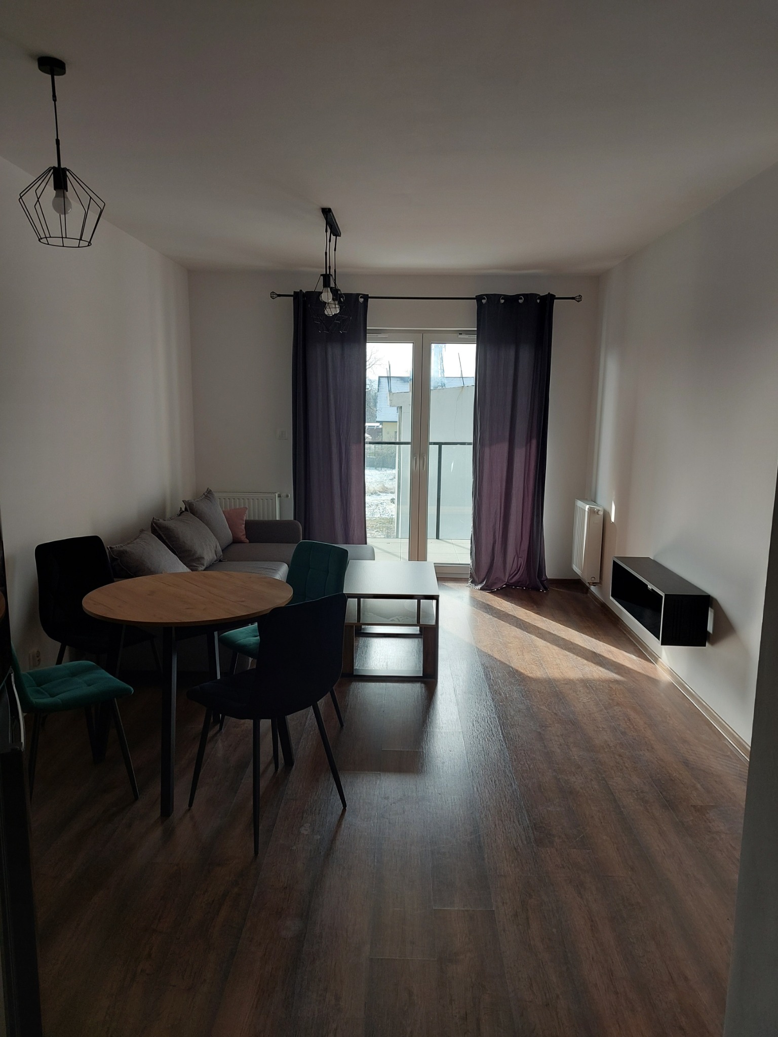 Jasne wnętrze salonu z drewnianą podłogą i meblami w stonowanych kolorach, oświetlone naturalnym światłem wpadającym przez okno balkonowe.