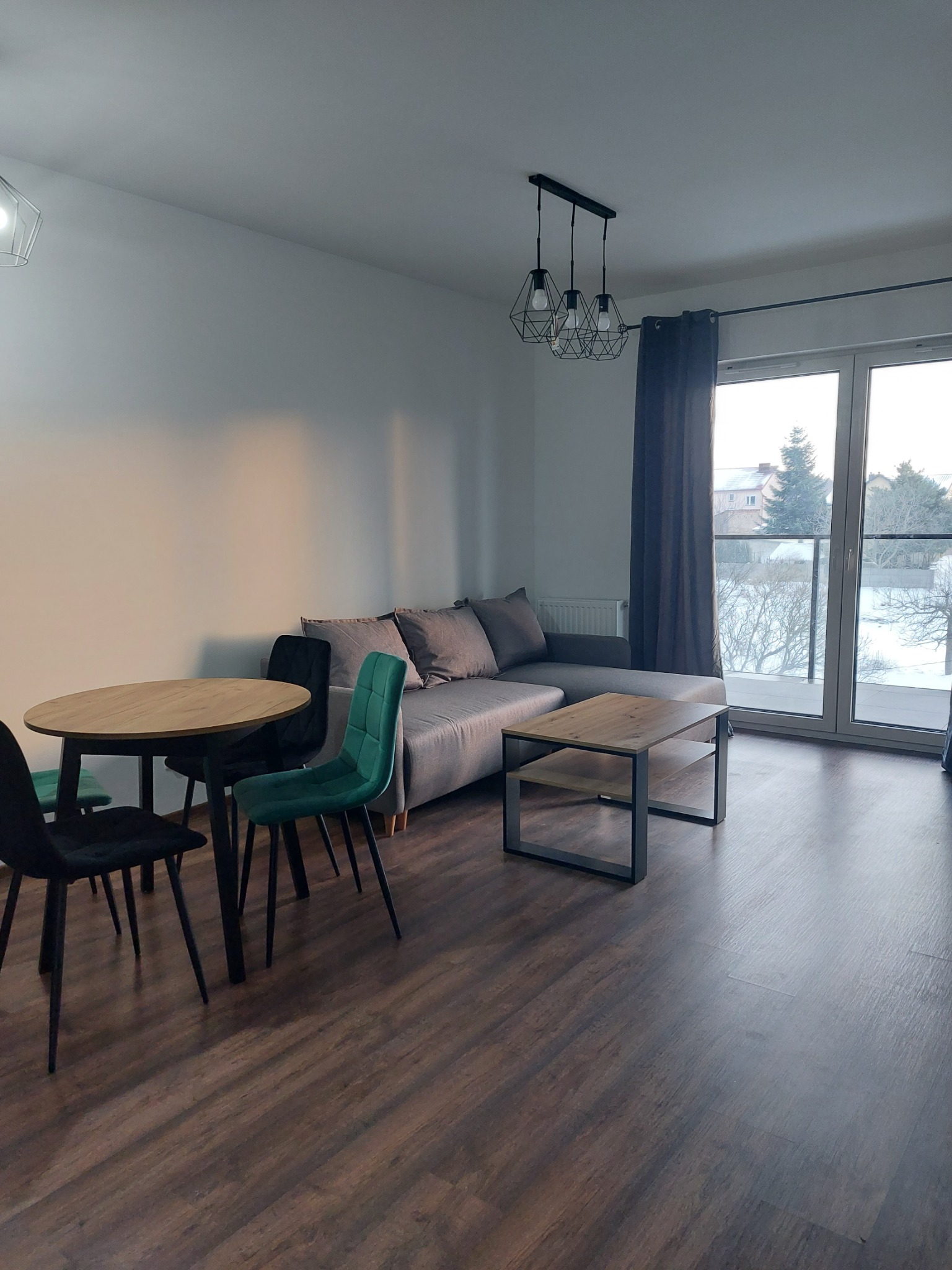 Nowoczesny salon z drewnianą podłogą, szarą sofą i okrągłym stołem. Widok na zasnieżony balkon przez duże okno. Minimalistyczny design i jasne wnętrze.