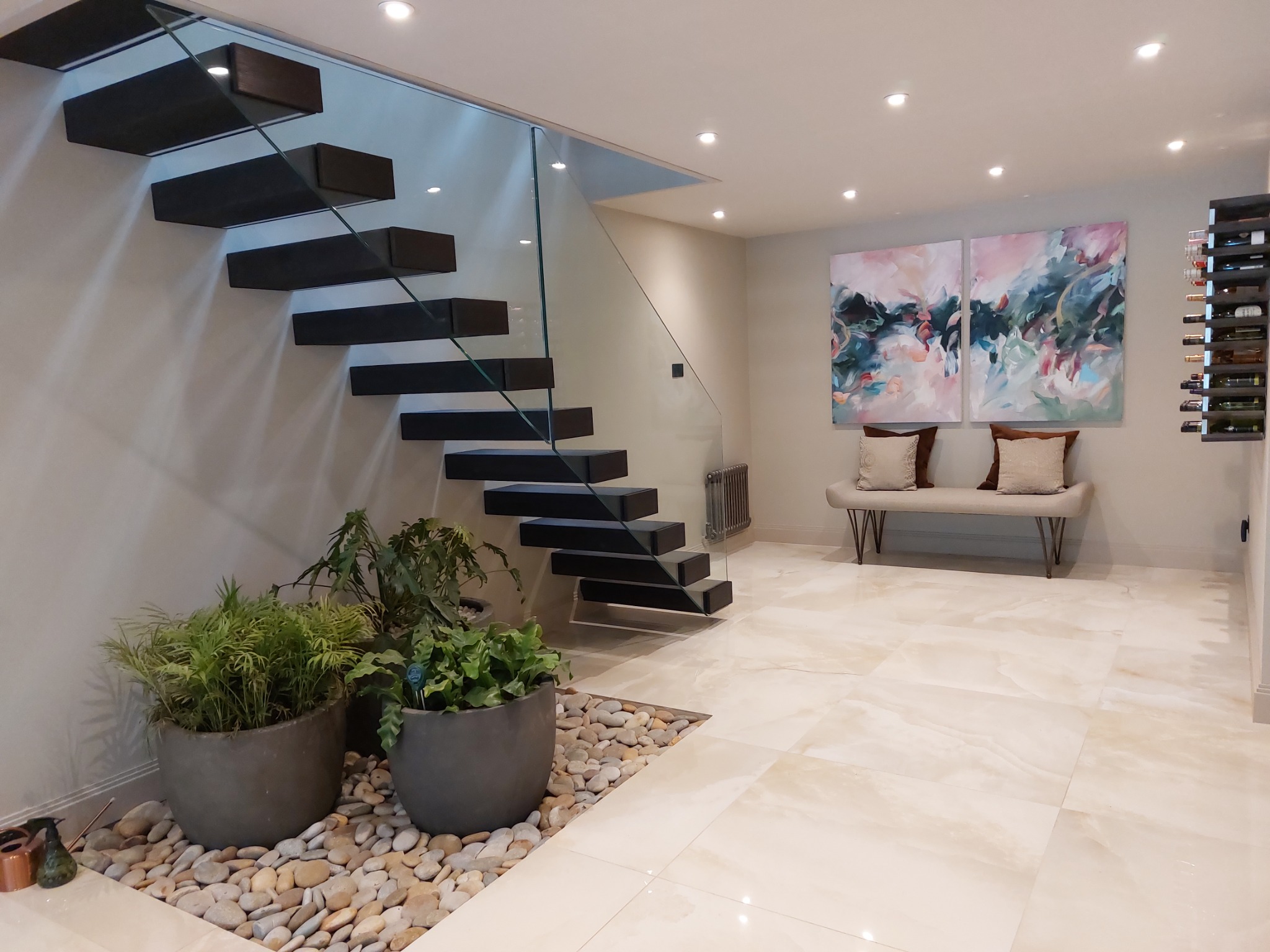 Nowoczesne schody wspornikowe z ciemnego drewna i szklaną balustradą, oświetlone punktowo, z roślinami doniczkowymi i jasną, marmurową podłogą.
