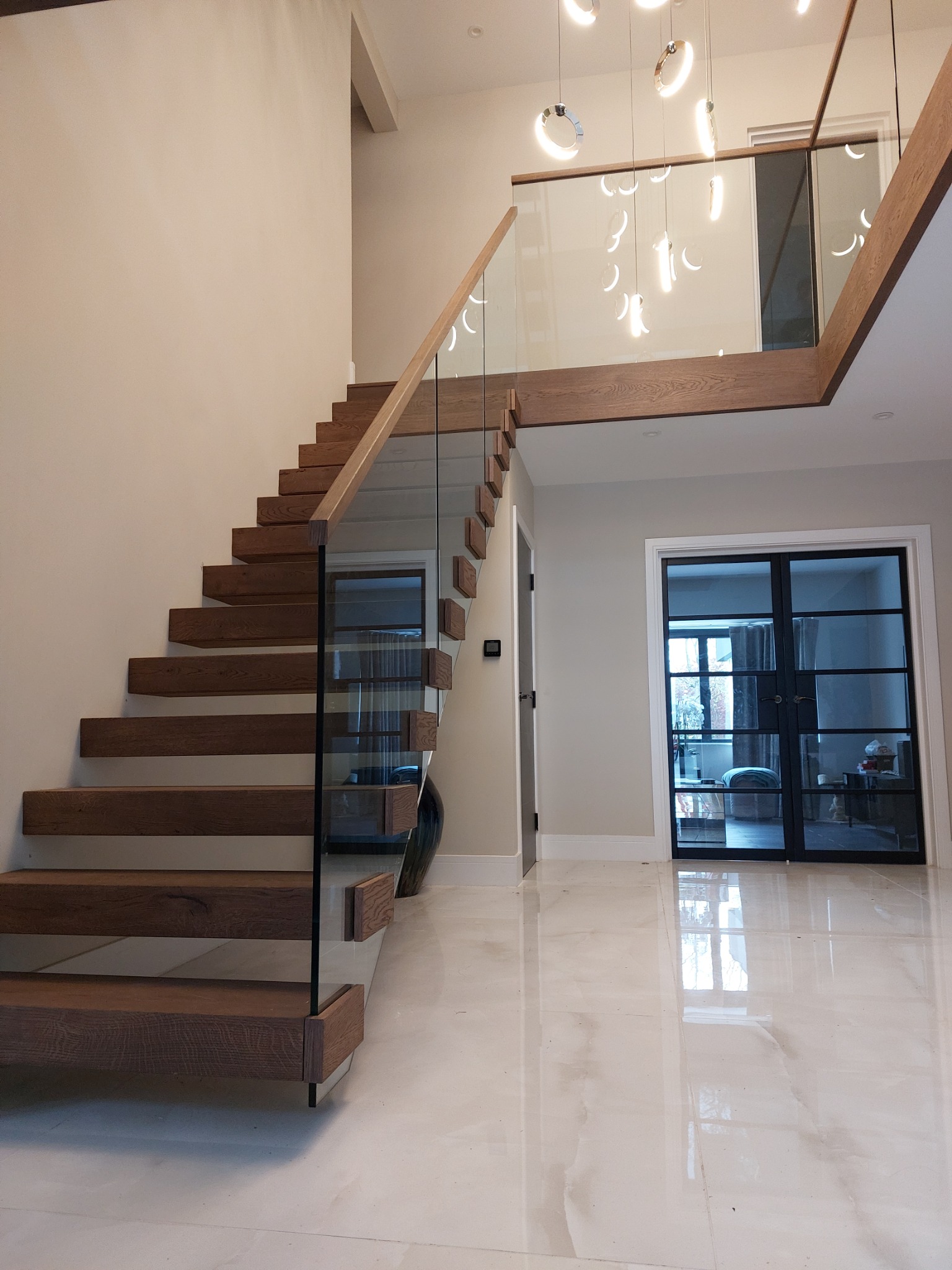 Nowoczesne, drewniane schody wspornikowe z szklaną balustradą w jasnym holu. Widoczne marmurowe płytki podłogowe i czarne, przeszklone drzwi.