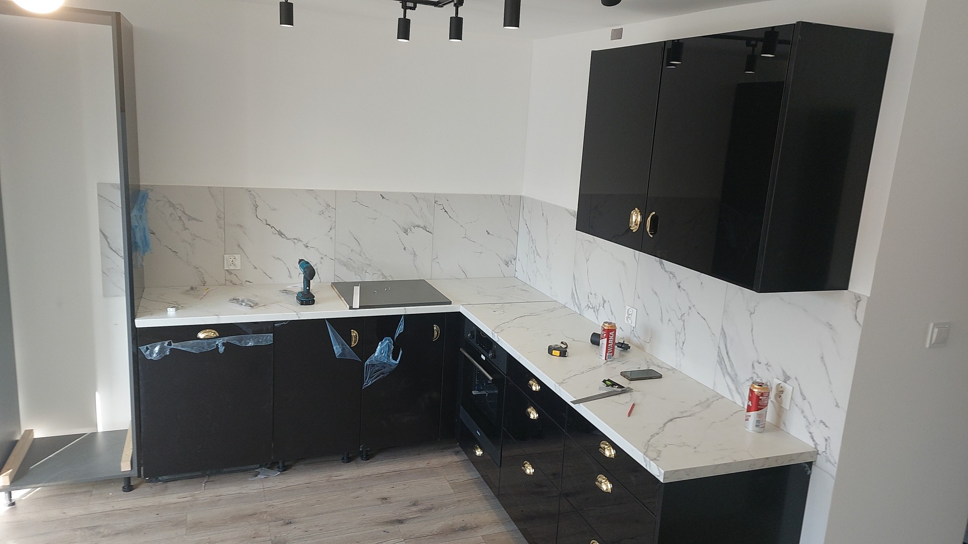 Nowoczesna, czarna kuchnia z marmurowymi płytkami na ścianie. Widoczny blat, szafki, narzędzia pracy i puszki. Ujęcie z perspektywy wnętrza, w trakcie montażu.