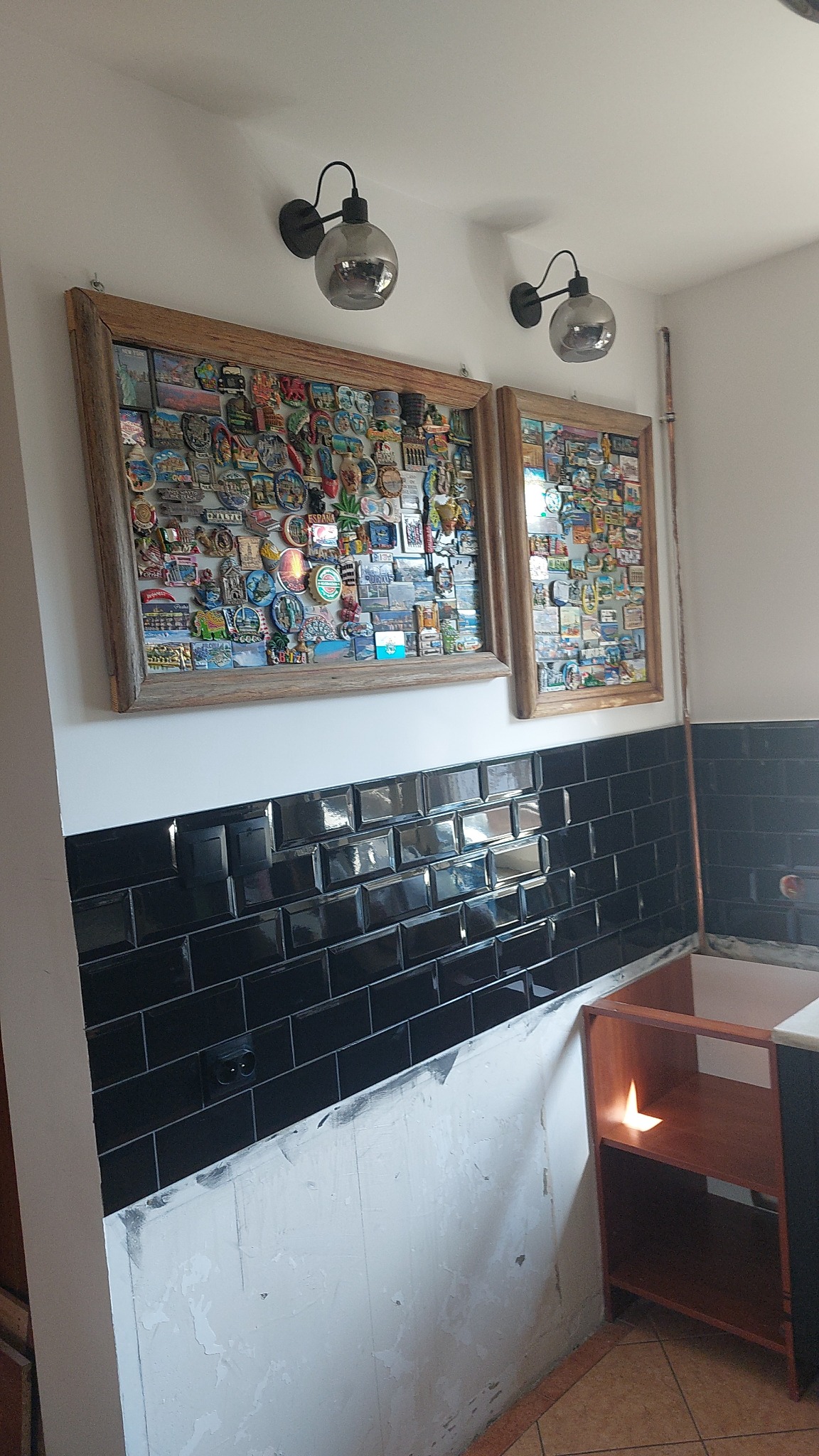 Czarne, błyszczące płytki ułożone na ścianie w kuchni, nad niezabezpieczoną ścianą. Nad nimi ramki z pamiątkowymi magnesami i lampy w stylu retro.