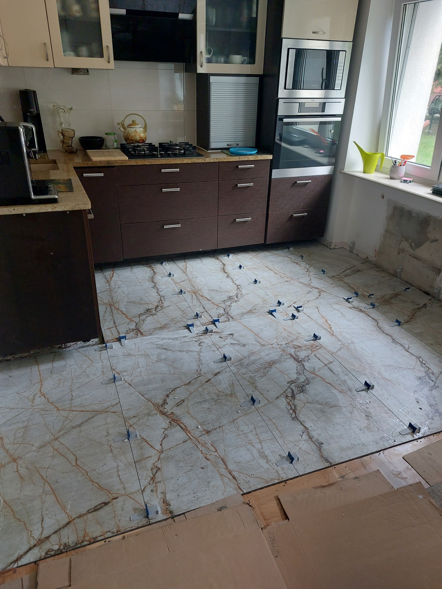 Kuchenna podłoga w trakcie układania płytek imitujących marmur, z użyciem niebieskich krzyżyków dystansowych, widoczne meble kuchenne i okno.