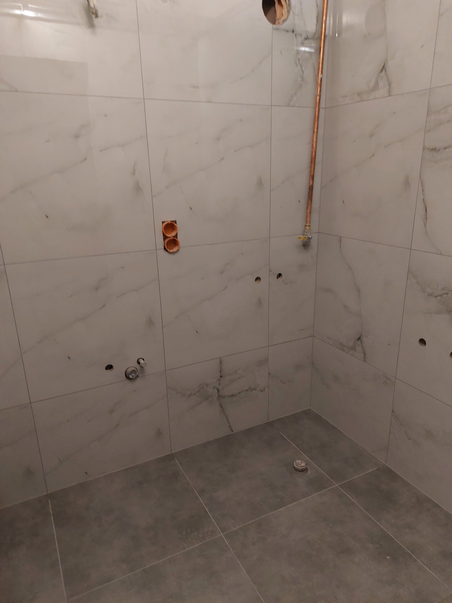 Wykończona łazienka z białymi marmurkowymi płytkami ściennymi i szarymi podłogowymi. Widoczne instalacje wodne i elektryczne przed montażem armatury.