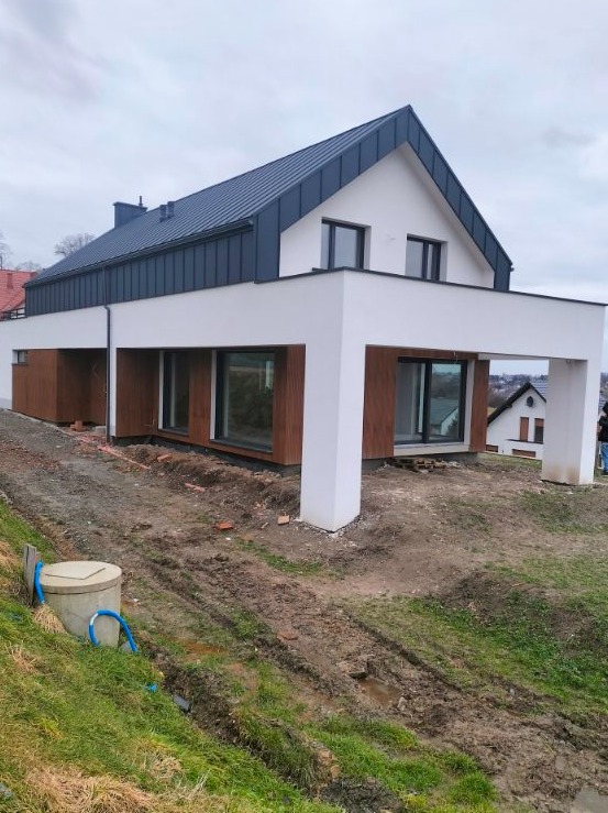 Nowoczesny dom jednorodzinny z białymi ścianami, grafitowym dachem i drewnianą elewacją w trakcie wykończenia, widoczne ślady prac ziemnych i betonowy zbiornik na pierwszym planie.