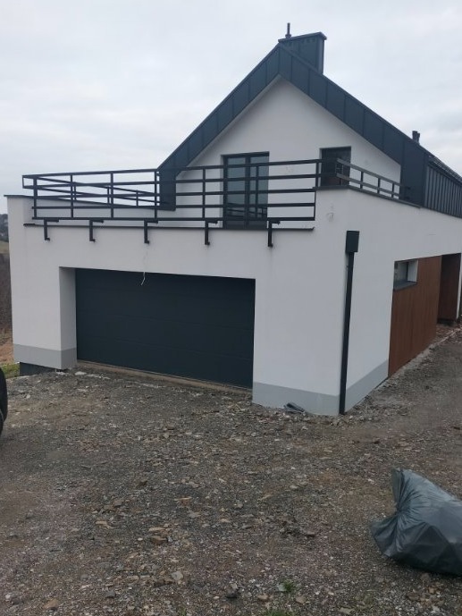 Nowoczesny dom z garażem i balkonem z czarną balustradą, świeżo po malowaniu elewacji na biało, z widocznym fragmentem brązowej boazerii i niezagospodarowanym terenem wokół.