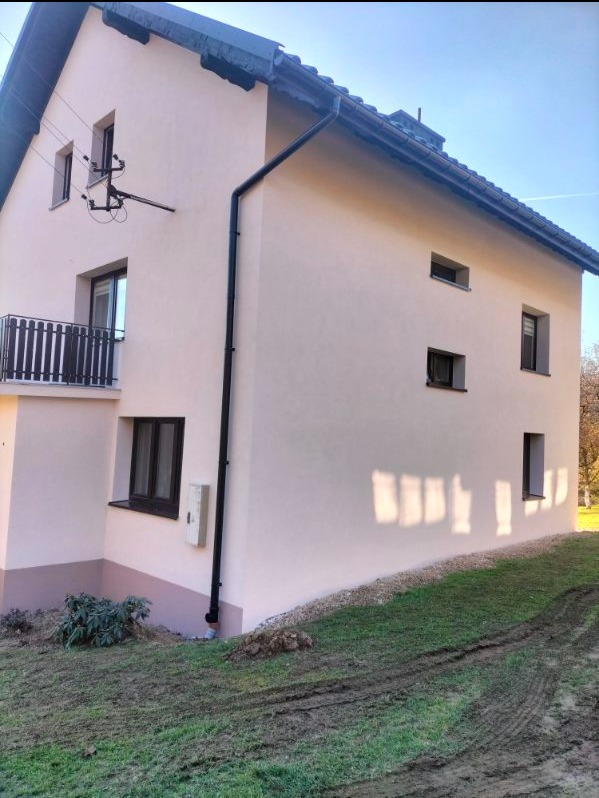 Dwupiętrowy dom z jasną elewacją i ciemnymi oknami, widok z boku, z widocznym balkonem i instalacją elektryczną na ścianie.