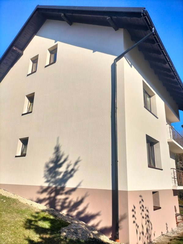 Dwukondygnacyjny dom z jasną elewacją i ciemnobrązowym pasem u dołu, widok z boku, z widocznym cieniem drzewa na ścianie i rynną.