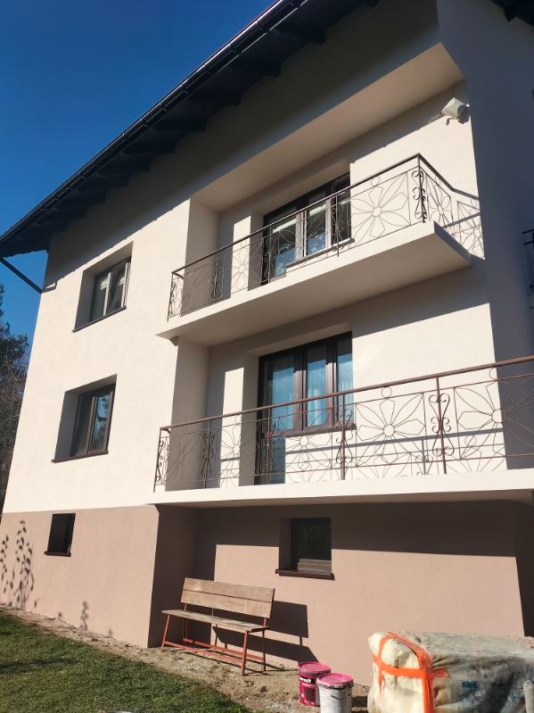 Dwupiętrowy dom z ocieploną elewacją w kolorze kremowym i brązowym, z balkonami z metalowymi balustradami o motywie kwiatowym, widoczne puszki z farbą i paczka materiału izolacyjnego.