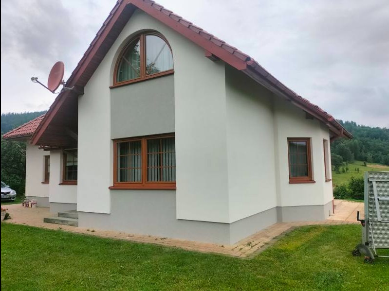 Dwukondygnacyjny dom jednorodzinny z ocieploną, białą elewacją i szarymi akcentami, brązowe okna z kratami, czerwona dachówka, satelita na dachu, zielony trawnik i górzyste tło.