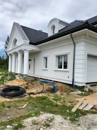 Nowoczesny dom jednorodzinny z białą elewacją, kolumnami przy wejściu i ciemnym dachem, widoczne wykopy wokół fundamentów z niebieskimi rurami i zwiniętym czarnym kablem.