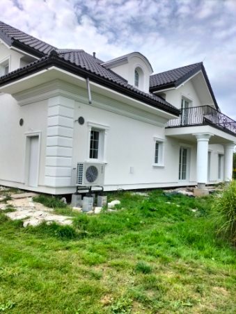 Biała elewacja domu jednorodzinnego z ciemną dachówką, widoczny klimatyzator na zewnątrz i fragment balkonu z kolumnami, trawnik z przodu.