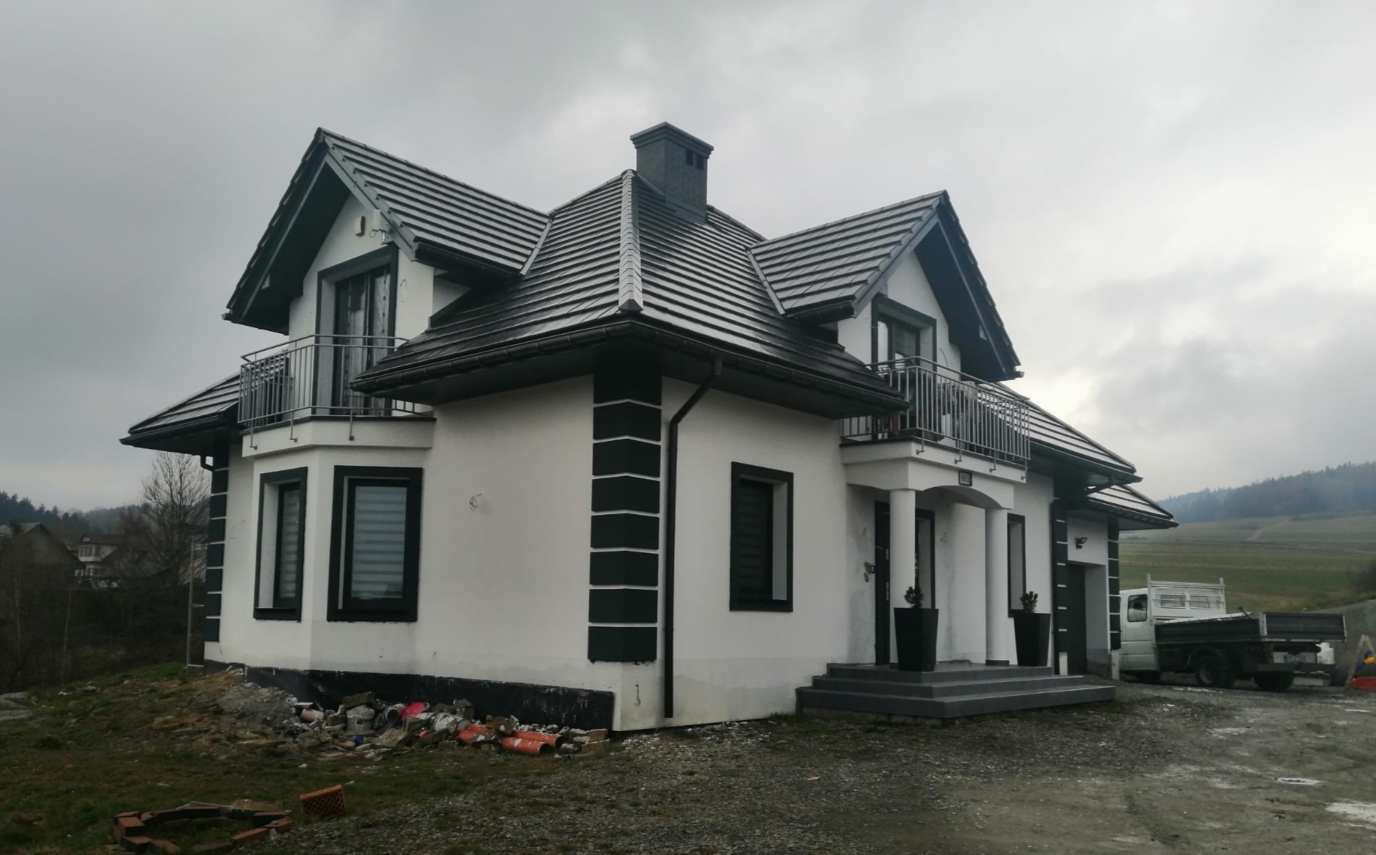 Nowoczesny dom jednorodzinny z białymi ścianami i ciemnymi detalami architektonicznymi, takimi jak obramowania okien, rynny i fragmenty elewacji, na tle pochmurnego nieba i krajobrazu zalesionych...