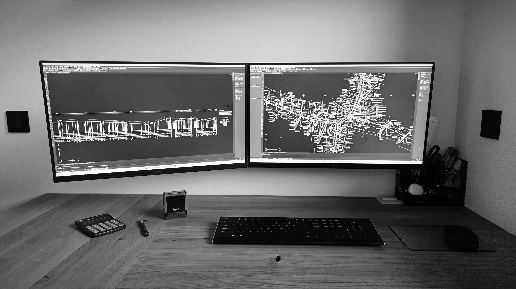 Dwa monitory MSI wyświetlają czarno-białe schematy instalacji sanitarnych na drewnianym biurku z kalkulatorem, stemplem Trodat i czarną klawiaturą.