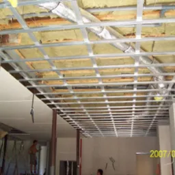 Montaż metalowej konstrukcji sufitu podwieszanego z widoczną izolacją i przewodami wentylacyjnymi, fragment ściany z cegły oraz pracownik na drabinie w tle.