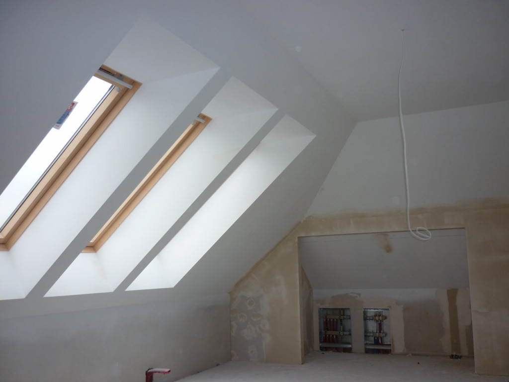 Poddasze w trakcie wykończenia z trzema oknami dachowymi, widoczne ślady po szpachlowaniu ścian i sufitu, oraz wystające przewody elektryczne.