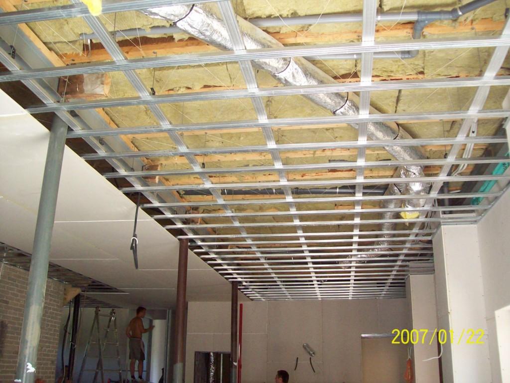 Montaż metalowej konstrukcji sufitu podwieszanego z widoczną izolacją i przewodami wentylacyjnymi, fragment ściany z cegły oraz pracownik na drabinie w tle.