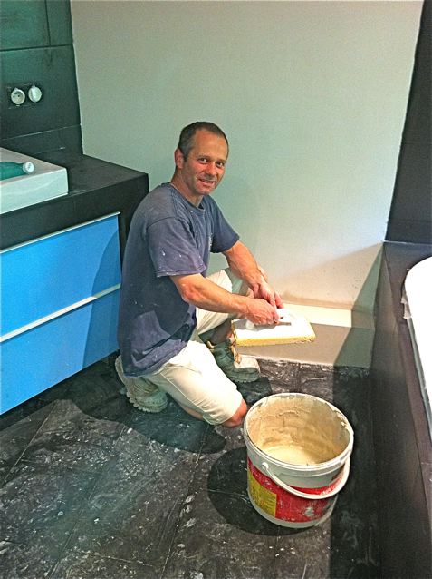 Mężczyzna w trakcie wygładzania szpachlą powierzchni w łazience z czarną podłogą i niebieską szafką pod umywalką, widoczny kubeł z masą szpachlową.