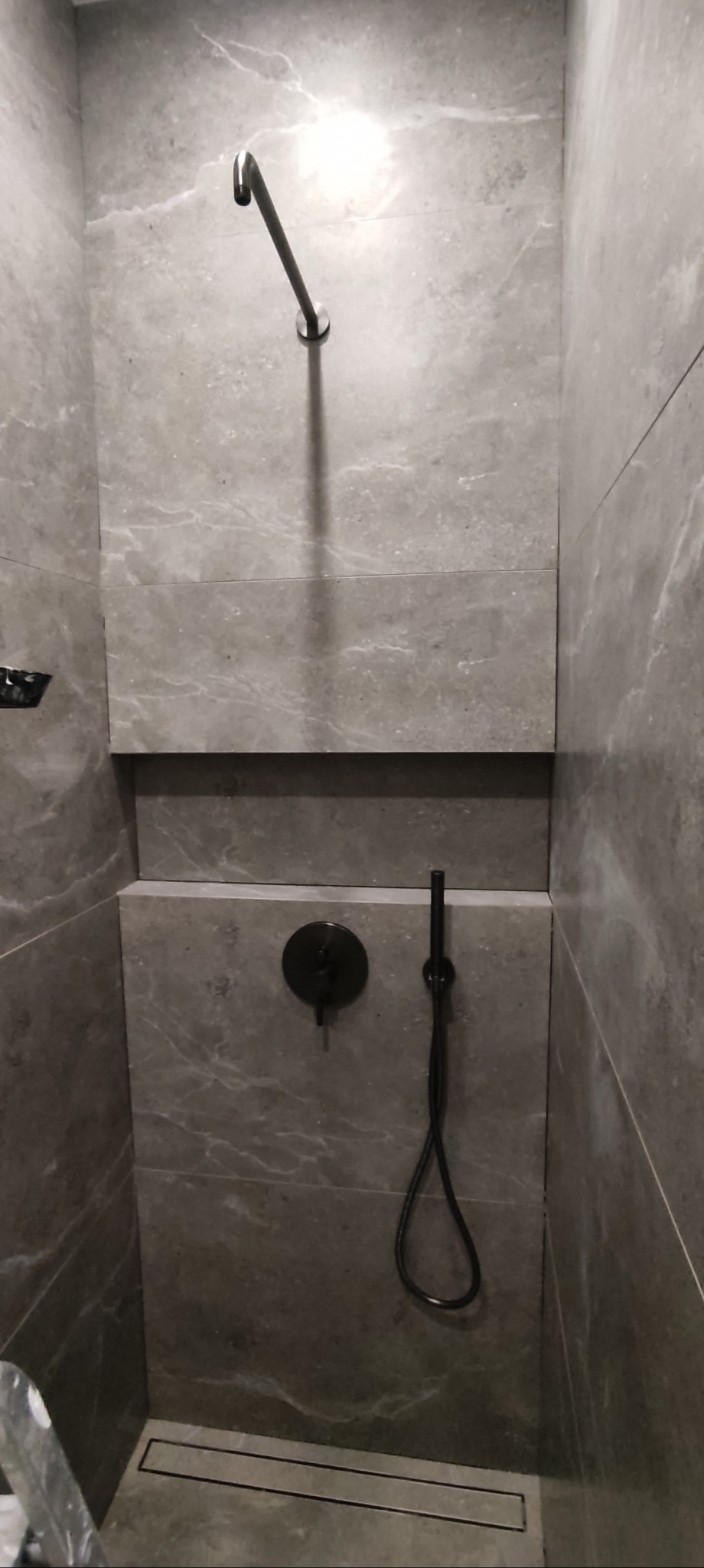 Szczegółowe ujęcie wykończonej kabiny prysznicowej z szarymi płytkami imitującymi marmur, czarną armaturą i odpływem liniowym w podłodze.