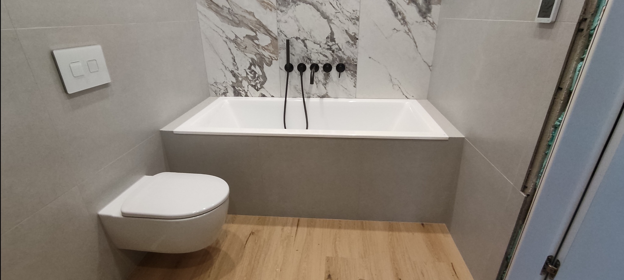 Nowoczesna łazienka z podwieszaną toaletą, białą wanną obudowaną szarą płytką i marmurową ścianą, czarna armatura, jasna drewnopodobna podłoga, widoczna puszka instalacyjna przy drzwiach.