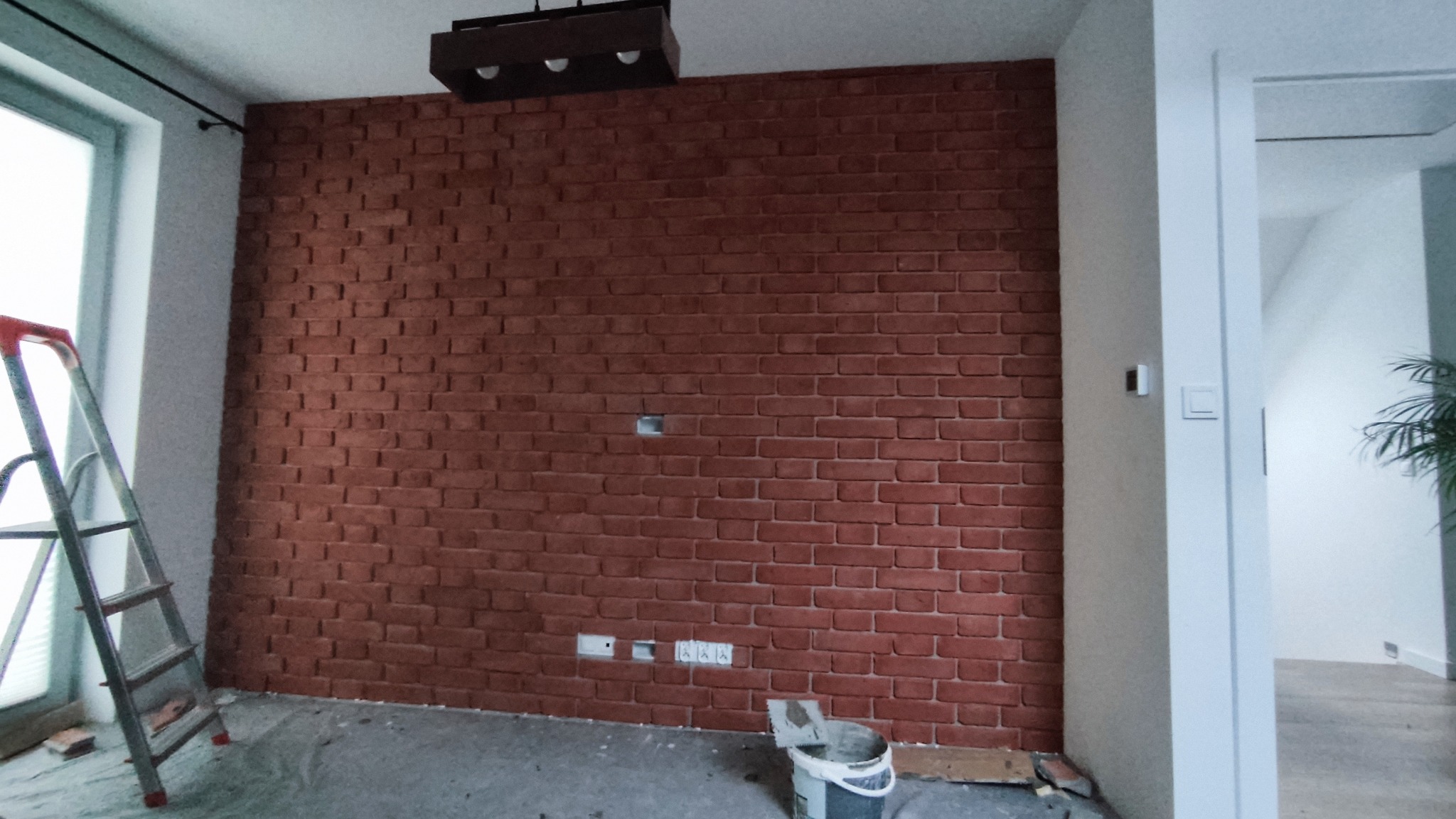 Czerwona ściana z cegły dekoracyjnej w trakcie montażu, widoczne puszki elektryczne, drabina oparta o ścianę, wiadro z zaprawą na podłodze zabezpieczonej folią.