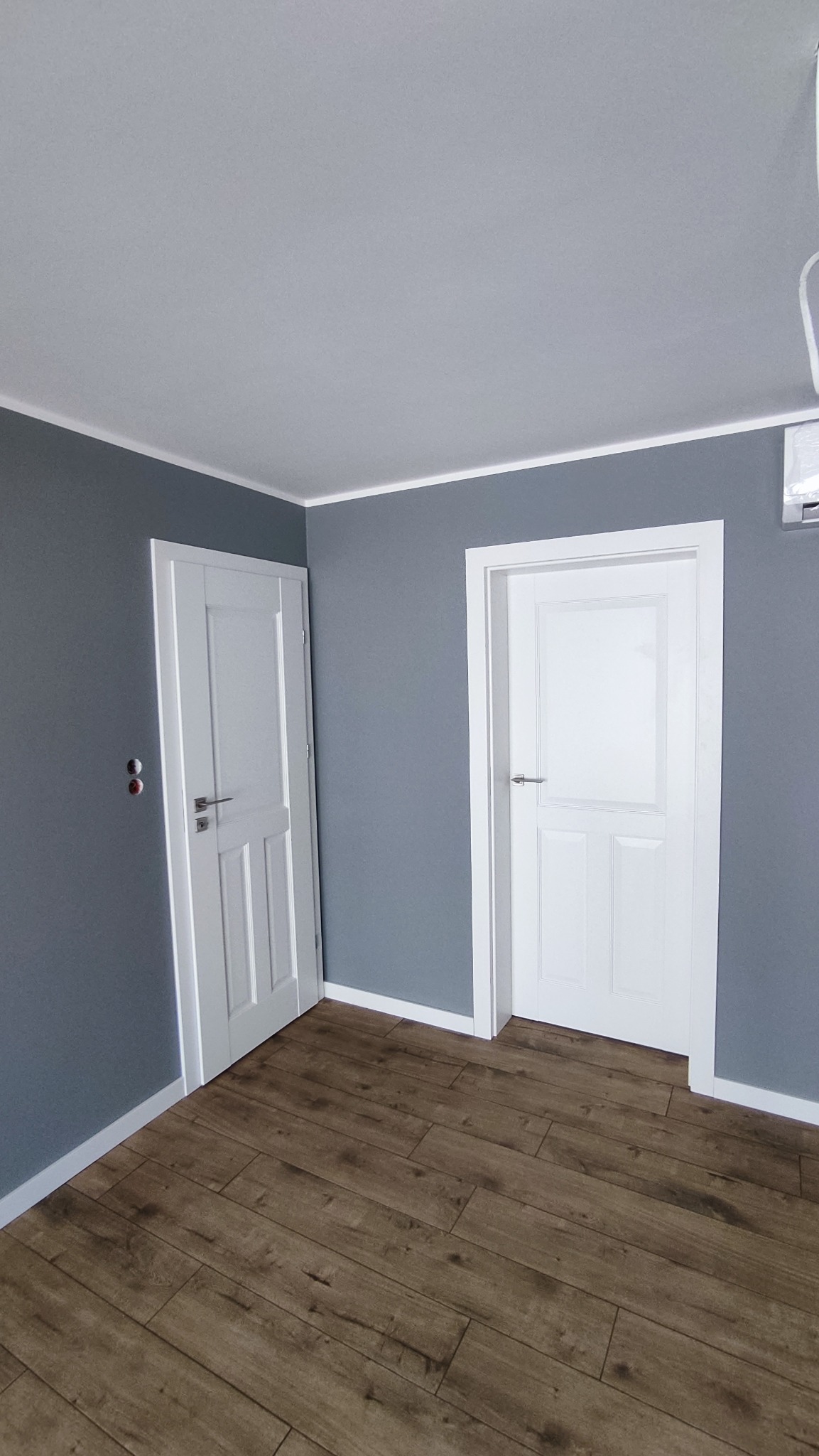 Pomieszczenie po remoncie: szare ściany, białe drzwi z panelami, podłoga z paneli imitujących drewno, listwy przypodłogowe.