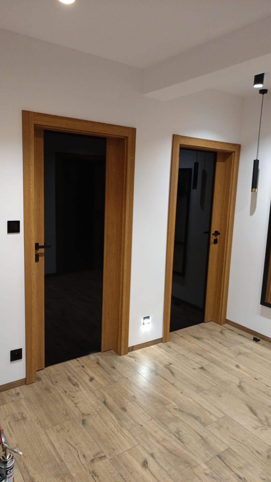 Dwa drewniane, wstawione drzwi z czarnym wypełnieniem, zamontowane w białej ścianie, widoczne oświetlenie punktowe i panele podłogowe imitujące drewno.