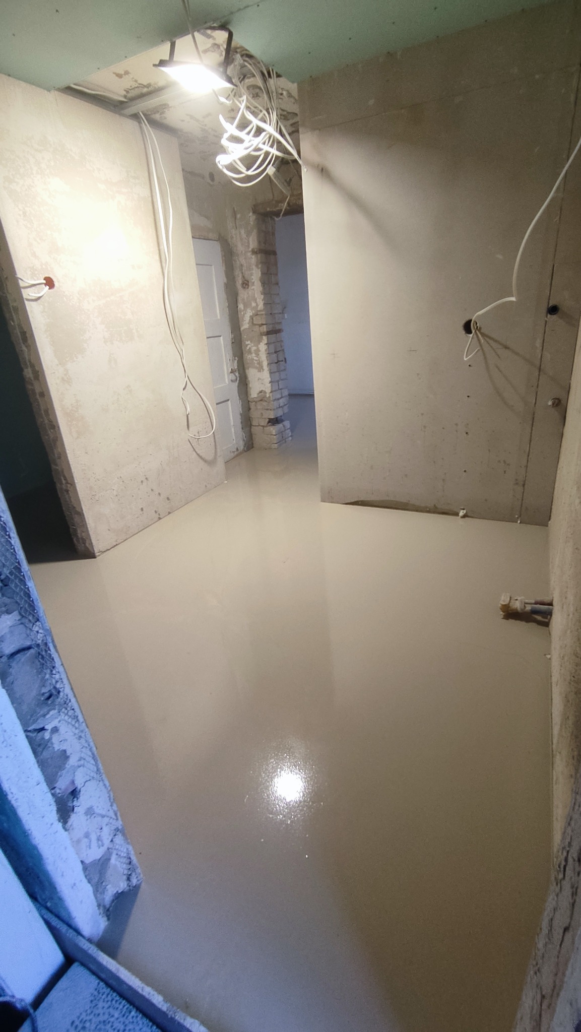 Świeżo wylana, samopoziomująca wylewka podłogowa w surowym wnętrzu, widoczne kable zwisające z sufitu i fragment ściany z cegły w przejściu do kolejnego pomieszczenia.