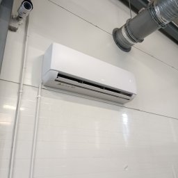 BART KLIMATYZACJA - Zamontowany klimatyzator AUX na białej ścianie z panelami, widoczne rury instalacyjne i kamera przemysłowa.