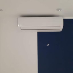 BART KLIMATYZACJA - Biała jednostka klimatyzacyjna zamontowana na ścianie biura z białymi i granatowymi panelami, widoczna lampa sufitowa.