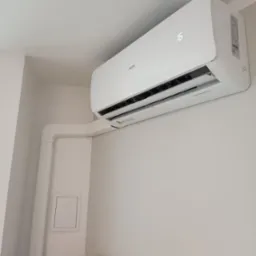 Biała jednostka klimatyzacyjna AUX zamontowana na ścianie w jasnym pomieszczeniu, widoczne białe rury instalacyjne i fragment mebla zabezpieczonego folią.