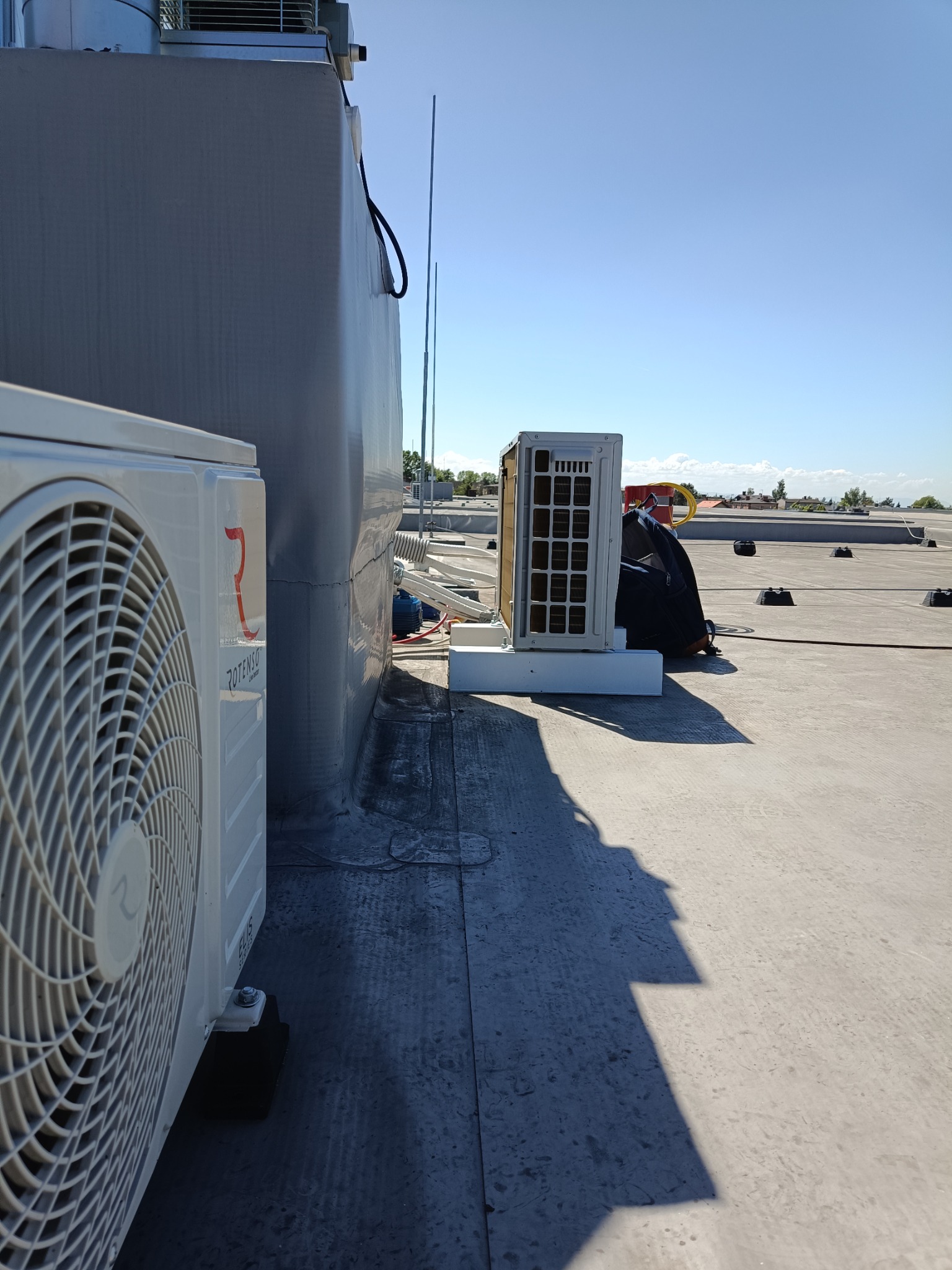 Dwa moduły klimatyzacji zamontowane na płaskim dachu budynku, jeden z widocznym logo 'Rotenso', w tle instalator w kombinezonie i narzędzia.