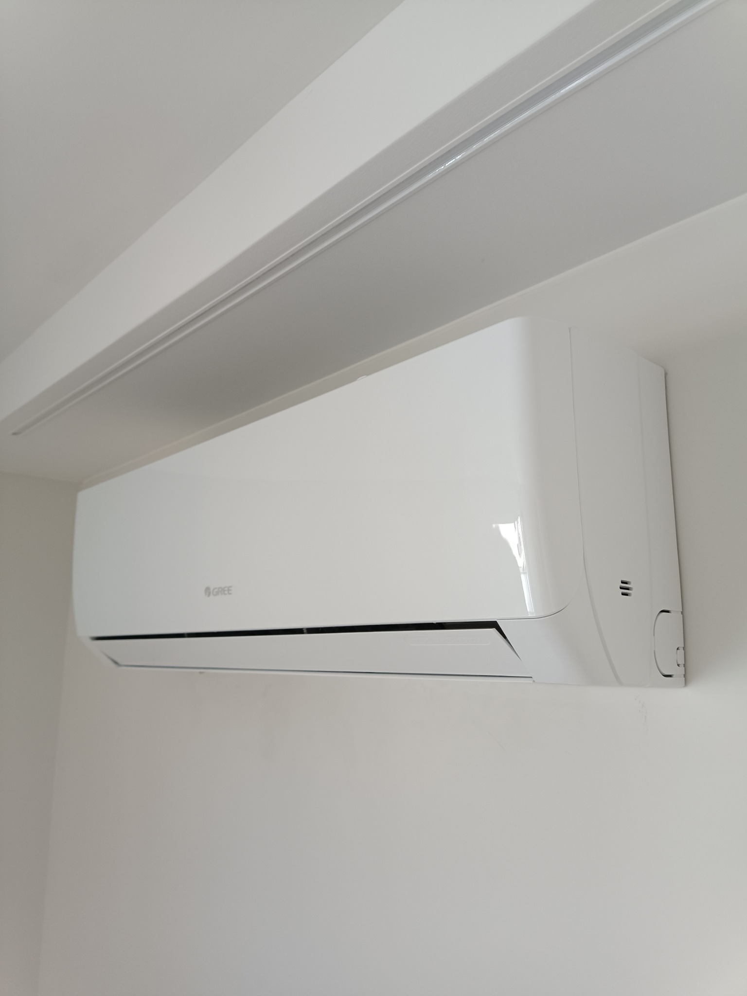 Biała jednostka klimatyzacji marki Gree zamontowana na jasnej ścianie w pomieszczeniu z minimalistycznym wykończeniem.