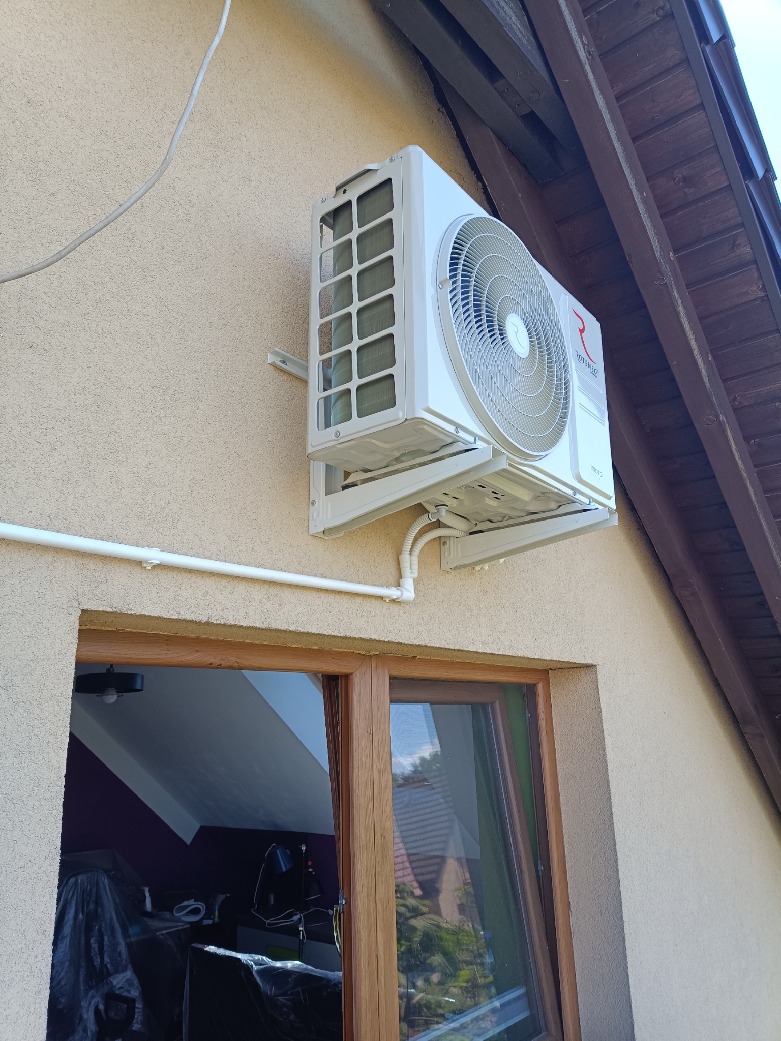 Biała jednostka zewnętrzna klimatyzacji Rotenso zamontowana na ścianie domu z beżowym tynkiem, widoczne rury instalacyjne i okno z drewnianą ramą.