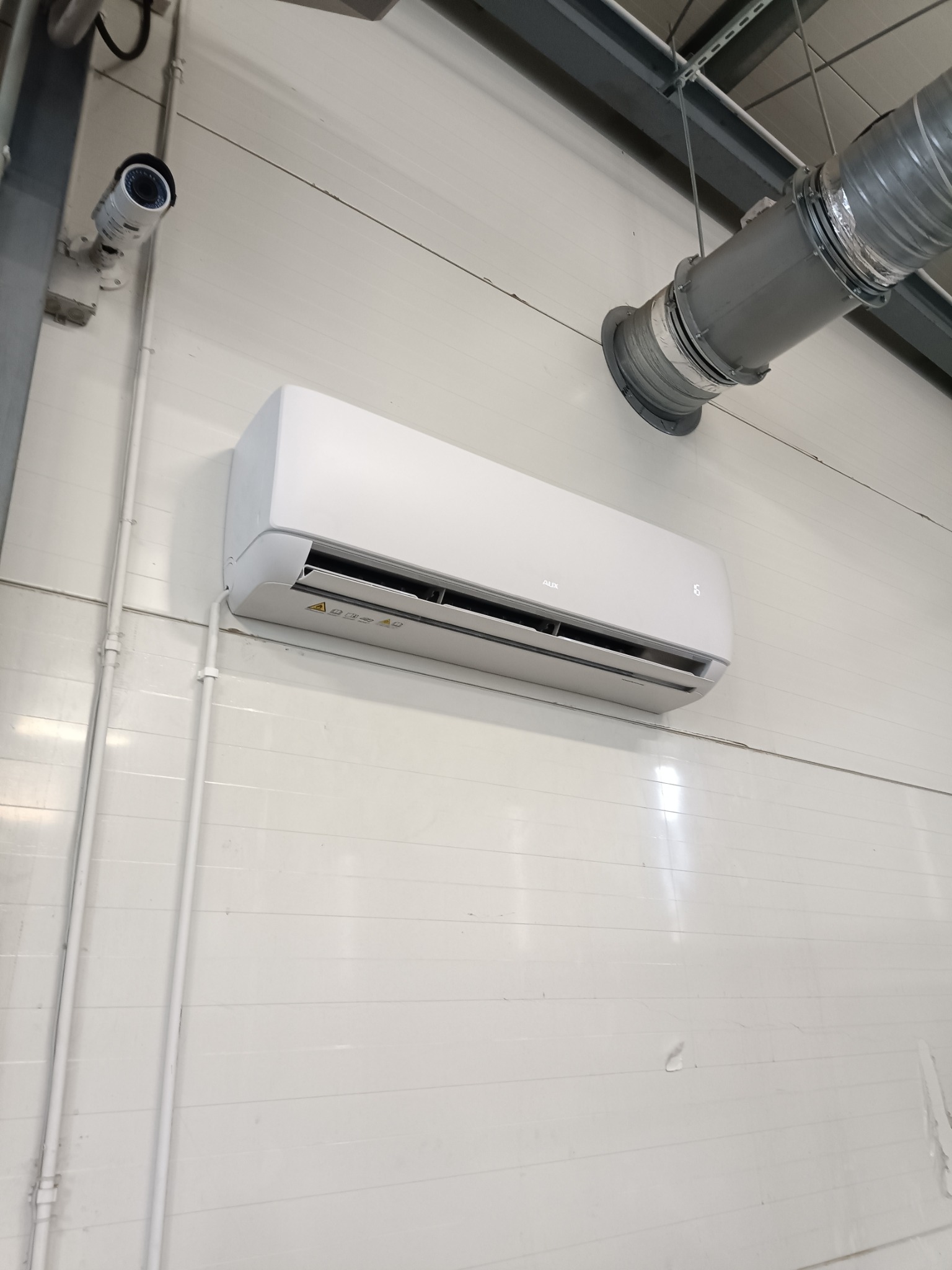 Zamontowany klimatyzator AUX na białej ścianie z panelami, widoczne rury instalacyjne i kamera przemysłowa.