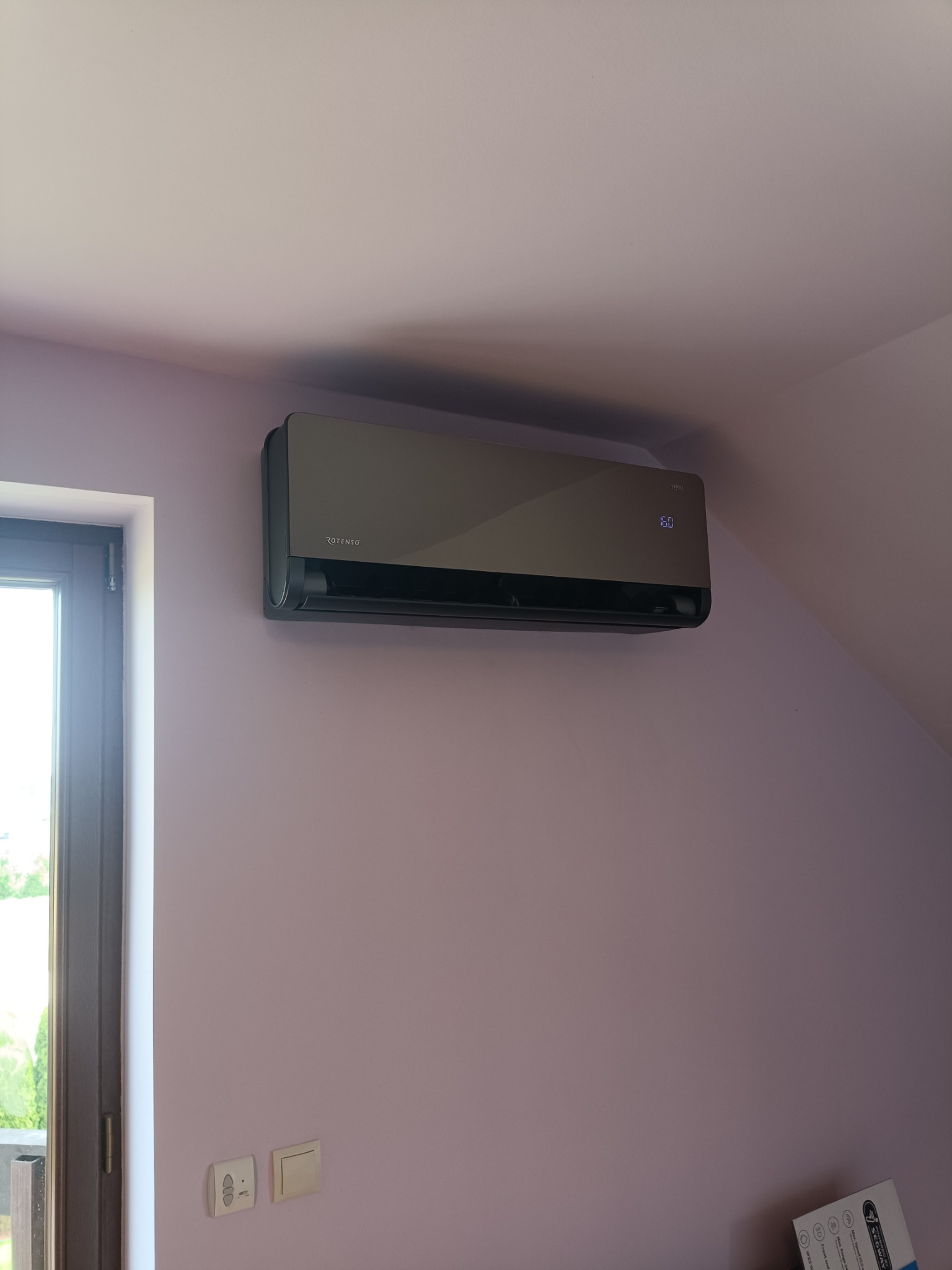 Szary klimatyzator marki Rotenso zamontowany na skośnej, fioletowej ścianie poddasza, obok okna balkonowego.