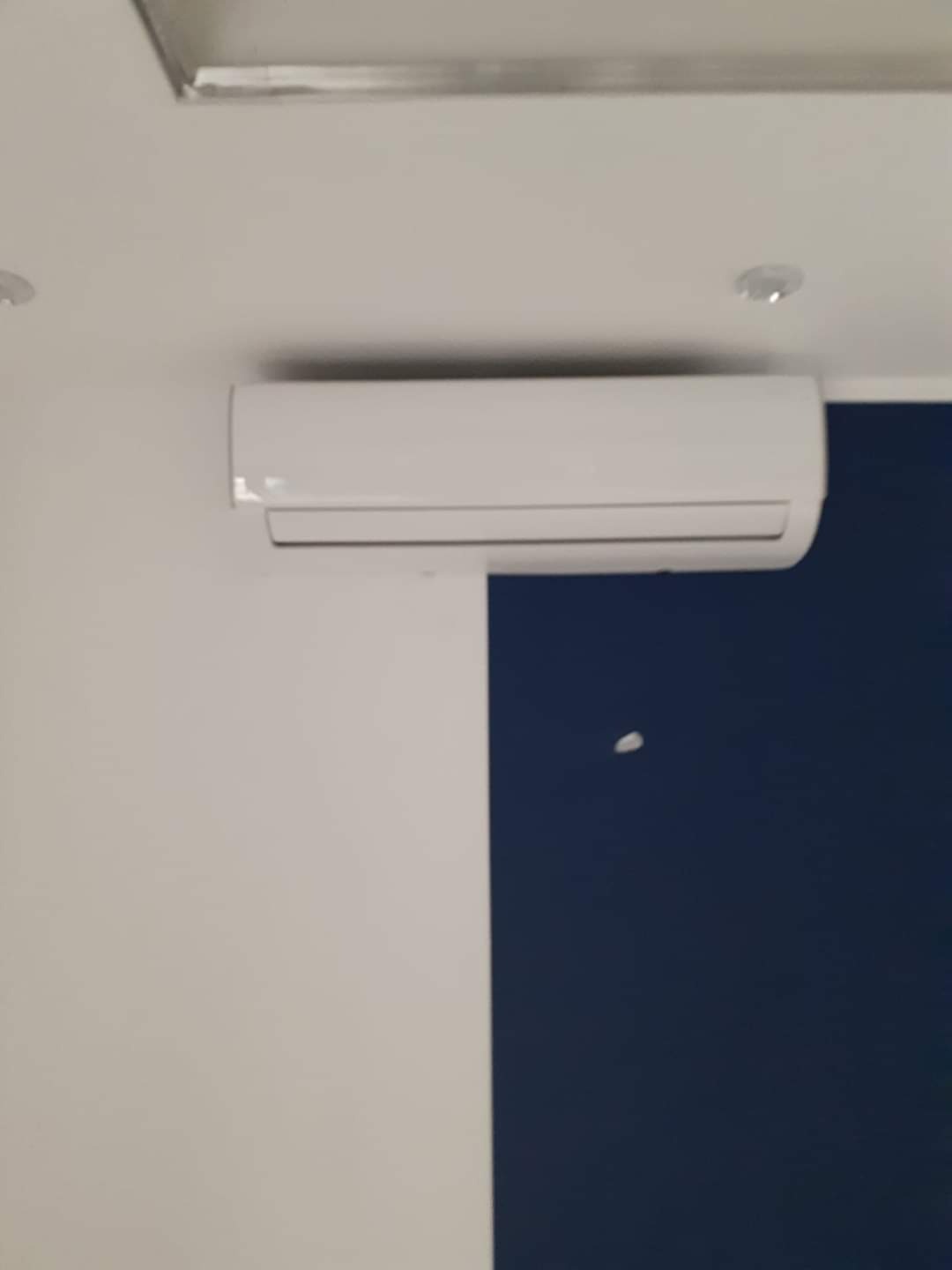 Biała jednostka klimatyzacyjna zamontowana na ścianie biura z białymi i granatowymi panelami, widoczna lampa sufitowa.