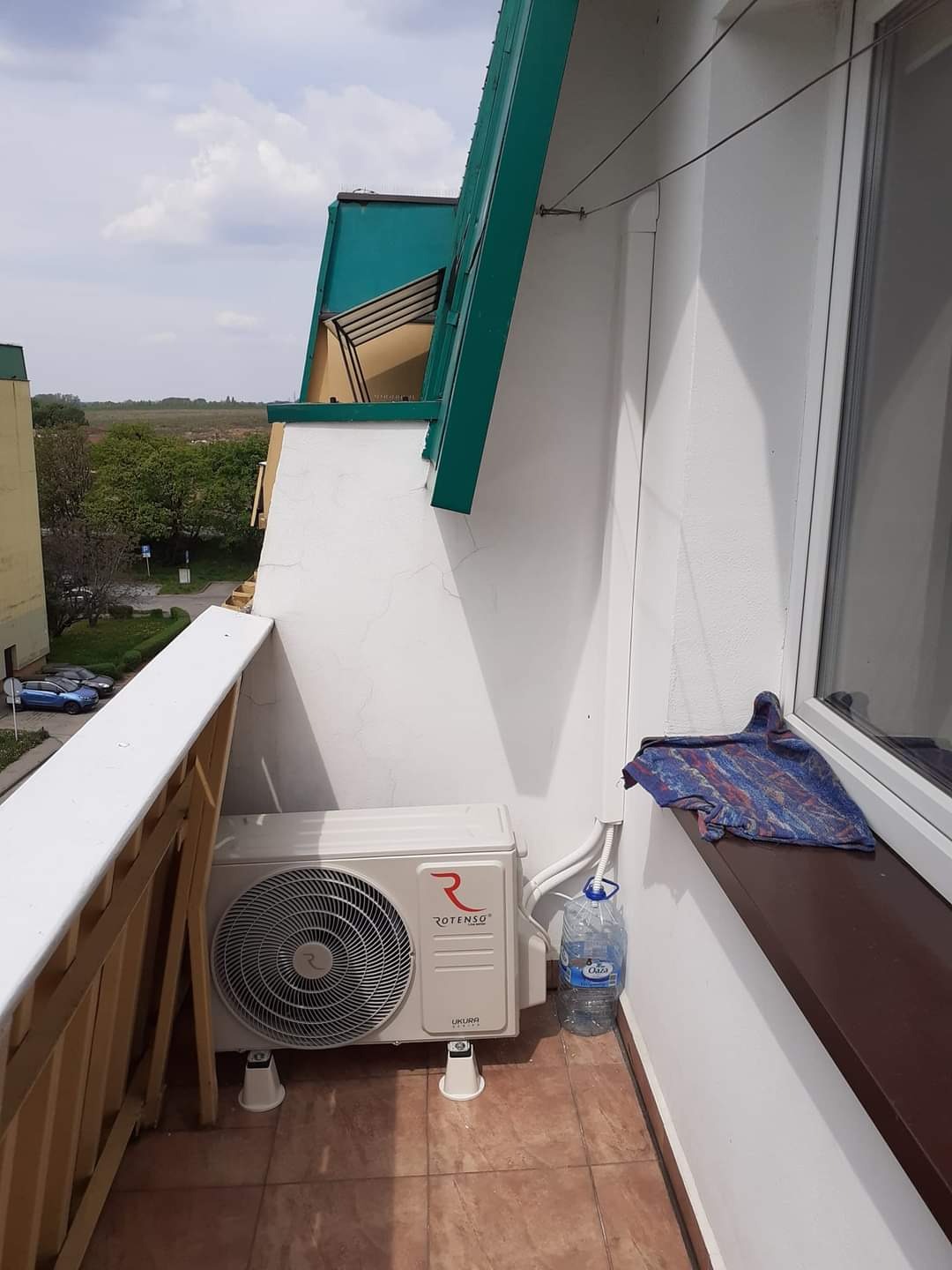 Jednostka zewnętrzna klimatyzacji Rotenso Ukura zamontowana na balkonie, widoczna butelka wody i szmatka na parapecie.