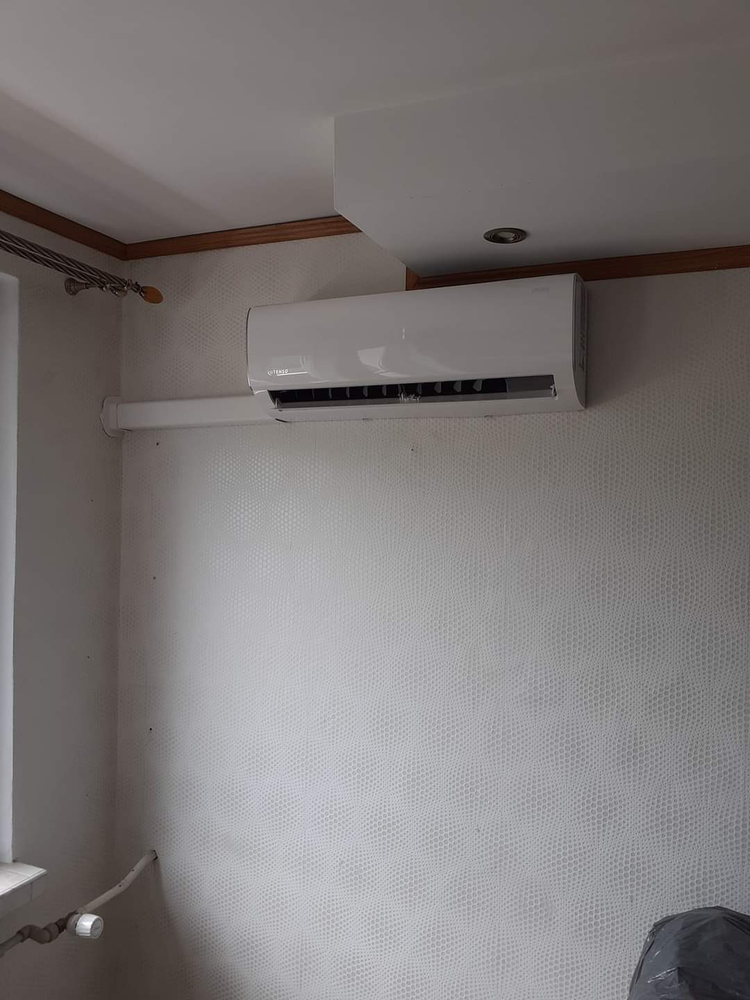 Biały klimatyzator ścienny marki Kotenso zamontowany w pokoju z jasną tapetą o wzorze drobnej siatki, widoczna instalacja odprowadzająca skropliny.