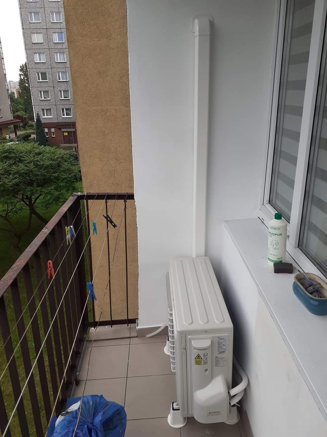 Biała jednostka zewnętrzna klimatyzacji zamontowana na balkonie mieszkania w bloku, widoczne rury odprowadzające skropliny i pionowa maskownica instalacji na ścianie, na balustradzie wiszą klamerki...