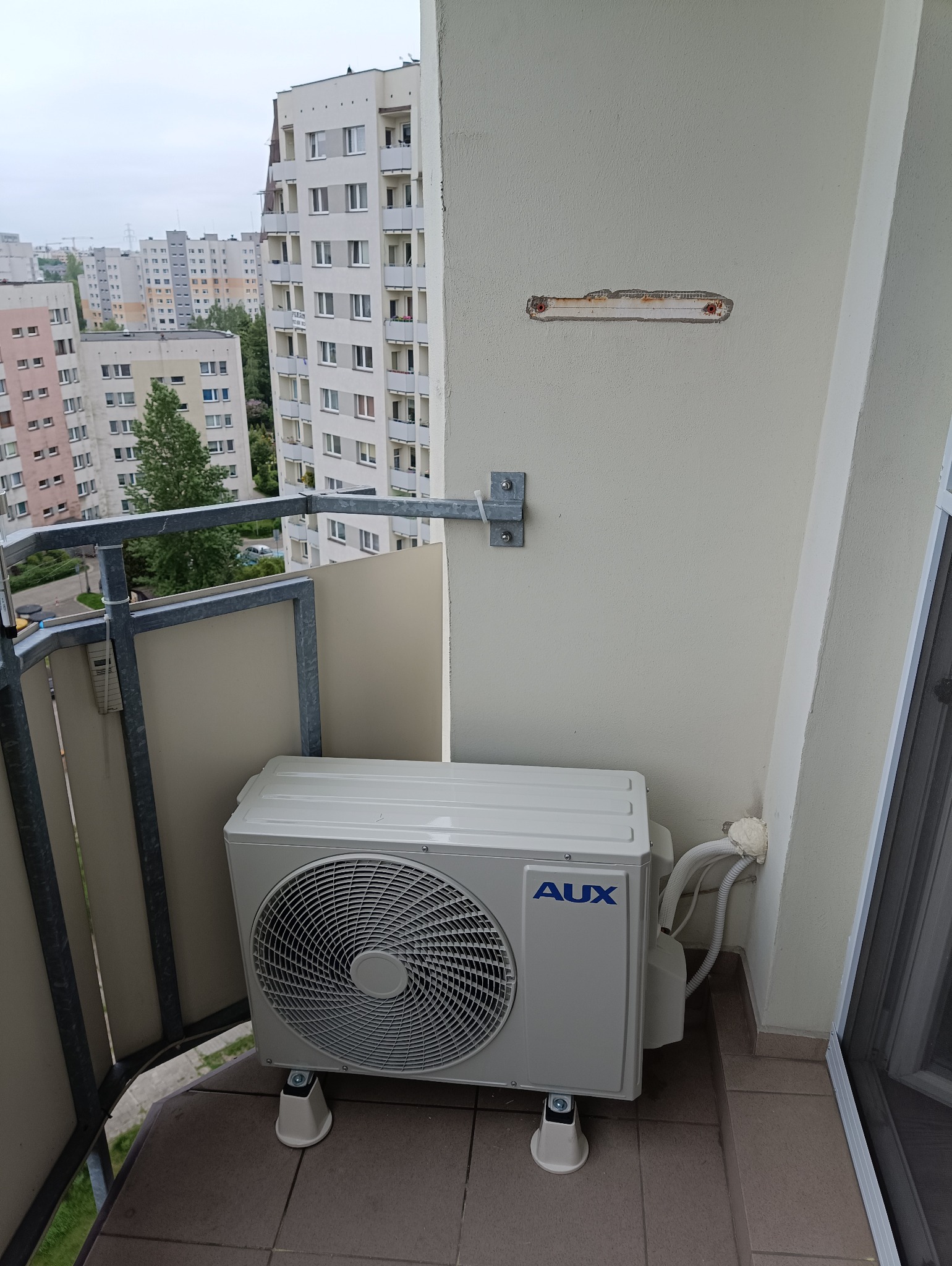 Zewnętrzna jednostka klimatyzacji marki AUX umieszczona na balkonie bloku mieszkalnego, widoczne przewody i mocowanie na ścianie, w tle zabudowa miejska.