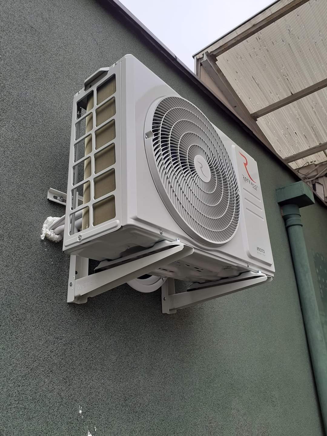 Biała jednostka zewnętrzna klimatyzacji marki Rotenso zamontowana na szarej, teksturowanej ścianie budynku z widocznym elementem zadaszenia.