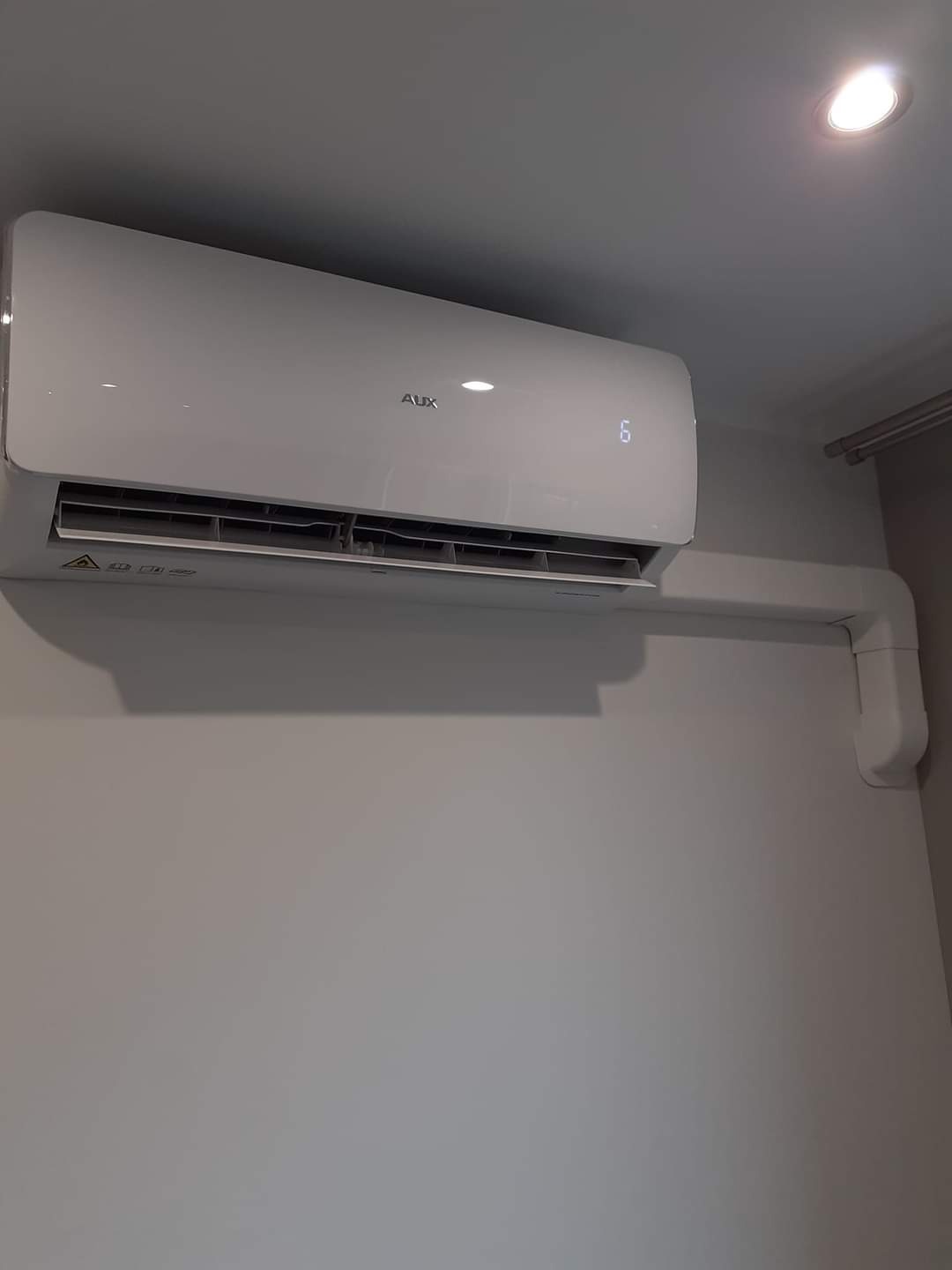 Biały klimatyzator marki AUX zamontowany na jasnej ścianie w pomieszczeniu, widoczne rury instalacyjne i wyświetlacz z cyfrą '6'.