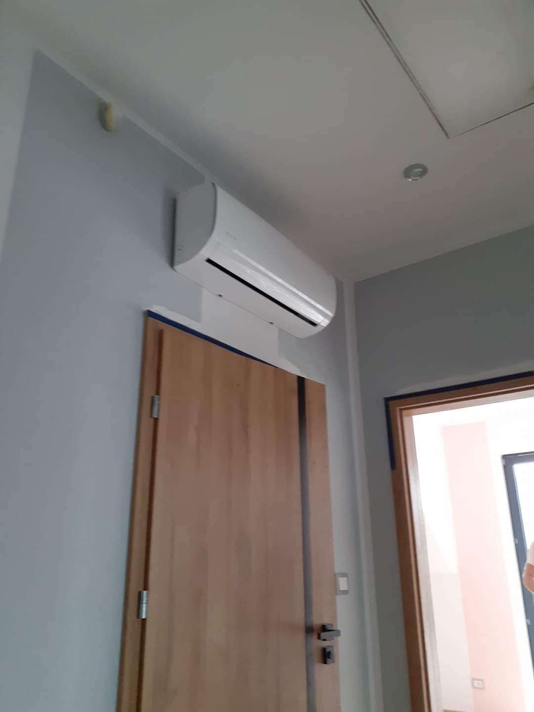 Biała jednostka klimatyzacyjna zamontowana nad drewnianymi drzwiami w jasnym pomieszczeniu, widok z dołu.